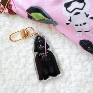 Pu&ograve; includere: Un portachiavi in acrilico trasparente con Darth Vader e una spada laser rosa. Il portachiavi ha un fermaglio color oro ed &egrave; posto su uno sfondo bianco morbido. Sullo sfondo &egrave; visibile un tessuto rosa con personaggi di Star Wars.