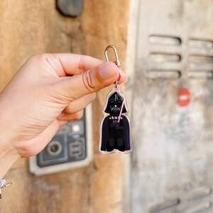 Pu&ograve; includere: Un portachiavi in acrilico trasparente con Darth Vader di Star Wars. Il portachiavi ha un fermaglio color oro e una spada laser rosa. La figura di Darth Vader &egrave; raffigurata in nero e grigio, con un elmo e un mantello dettagliati.