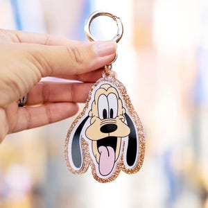 May include: A Pluto character keychain with a gold-colored clasp and glittery rose gold border. The cartoon dog has a tan face, black ears, and a pink tongue. The keychain is held in a person's hand.