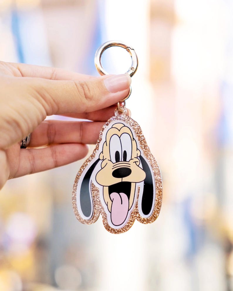 May include: A Pluto character keychain with a gold-colored clasp and glittery rose gold border. The cartoon dog has a tan face, black ears, and a pink tongue. The keychain is held in a person's hand.