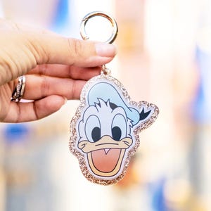 May include: A Donald Duck keychain with a gold-colored clasp. The keychain features a cartoon image of Donald Duck with a light blue hat and a glittery gold border. The background is blurred.