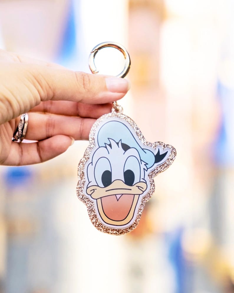 May include: A Donald Duck keychain with a gold-colored clasp. The keychain features a cartoon image of Donald Duck with a light blue hat and a glittery gold border. The background is blurred.