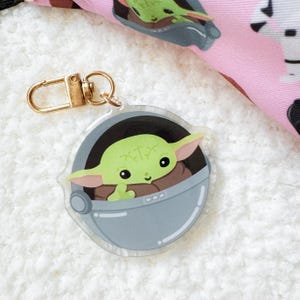 Pu&ograve; includere: Un portachiavi in acrilico trasparente con un personaggio dei cartoni animati verde in una capsula grigia. Il portachiavi ha una chiusura color oro. Lo sfondo &egrave; una superficie bianca morbida con un tessuto a fantasia rosa e nero.