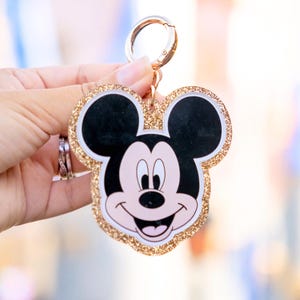 May include: A Mickey Mouse-shaped keychain with a gold glitter border. The keychain features a smiling Mickey Mouse face with black ears, a pink face, and a white mouth. The keychain has a gold-colored clasp.