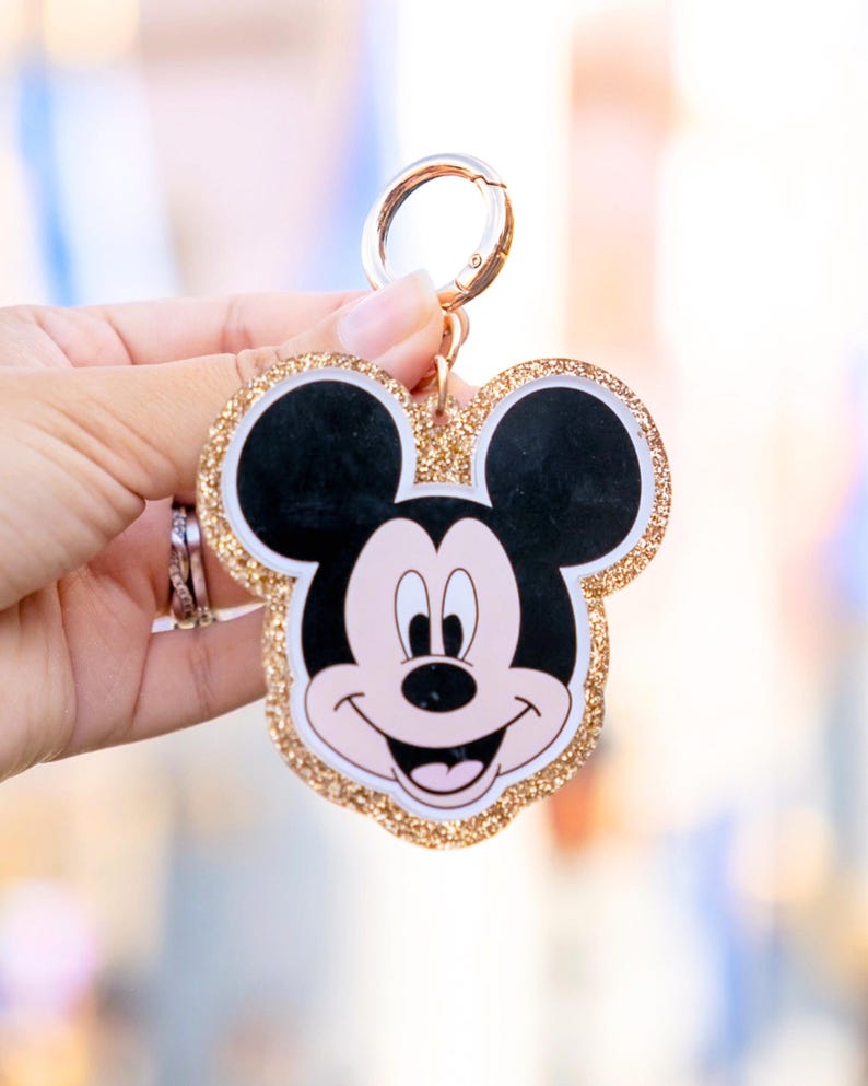 May include: A Mickey Mouse-shaped keychain with a gold glitter border. The keychain features a smiling Mickey Mouse face with black ears, a pink face, and a white mouth. The keychain has a gold-colored clasp.