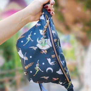 Può includere: Marsupio blu navy con un motivo ripetuto di Peter Pan e altri personaggi. Ha una cerniera dorata e un ciondolo di Peter Pan. Un accessorio pratico ed elegante per le tue avventure.