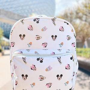 May include: White backpack featuring a repeating pattern of Mickey Mouse, Minnie Mouse, Donald Duck, and Daisy Duck faces. The backpack has a front pocket with a white zipper and a top handle.