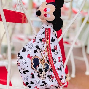 May include: A white and red Mickey Mouse-themed sling bag with a gold zip and a Mickey Mouse plushie attached. The bag features a pattern of Mickey Mouse, Minnie Mouse, and red bows. A heart-shaped keyring with Mickey Mouse is also attached.