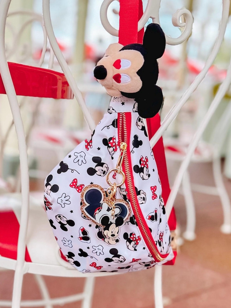 May include: A white and red Mickey Mouse-themed sling bag with a gold zip and a Mickey Mouse plushie attached. The bag features a pattern of Mickey Mouse, Minnie Mouse, and red bows. A heart-shaped keyring with Mickey Mouse is also attached.