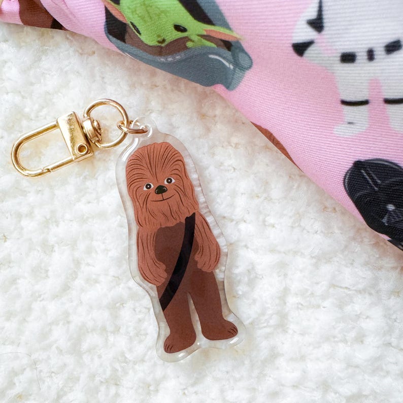 Pu&ograve; includere: Un portachiavi in acrilico trasparente con un personaggio dei cartoni animati di Chewbacca con pelliccia marrone e una cinghia nera. Il portachiavi ha una chiusura color oro. Un tessuto rosa con personaggi di Star Wars &egrave; sullo sfondo.