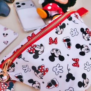May include: White fanny pack with a red zip and gold hardware, patterned with Mickey and Minnie Mouse illustrations and red polka dot bows. A Minnie Mouse plush toy and phone are also visible.