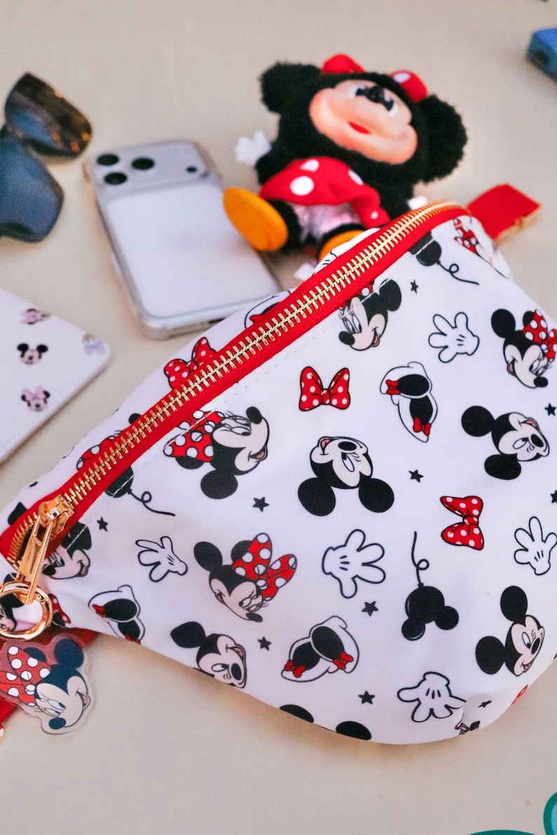 May include: White fanny pack with a red zip and gold hardware, patterned with Mickey and Minnie Mouse illustrations and red polka dot bows. A Minnie Mouse plush toy and phone are also visible.