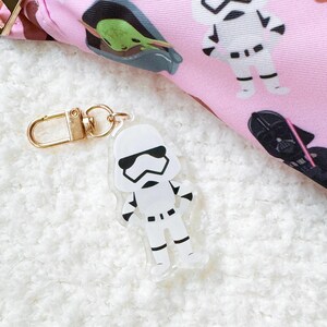 Pu&ograve; includere: Un portachiavi in acrilico trasparente con un personaggio di Stormtrooper bianco con dettagli neri. Il portachiavi ha un fermaglio color oro. Lo sfondo include un tessuto rosa con disegni di personaggi di Star Wars.