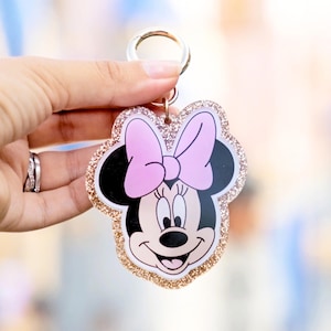 May include: A Minnie Mouse keychain with a pink bow and gold glitter trim. The keychain features a smiling Minnie Mouse face with black ears and a pink bow. The keychain is attached to a silver metal ring.