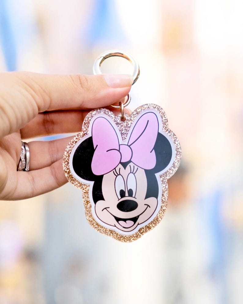 May include: A Minnie Mouse keychain with a pink bow and gold glitter trim. The keychain features a smiling Minnie Mouse face with black ears and a pink bow. The keychain is attached to a silver metal ring.