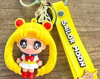 Op de afbeelding: Een gele Sailor Moon sleutelhanger met een cartoonfiguur met geel haar en een rode strik. De sleutelhanger bevat een gele band met "SAILOR MOON" in zwarte tekst en een goudkleurige sluiting. De tekst "Vimo's Gift Shop" staat onderaan.