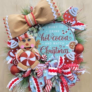 May include: A festive Christmas wreath featuring a gingerbread man, a large tan bow, and red and white striped ribbons. The wreath includes a sign that reads "I want to drink hot cocoa and watch Christmas."