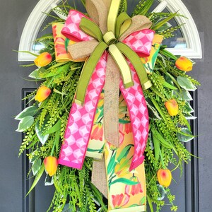 May include: A spring wreath with a burlap and green bow, accented with pink and white patterned ribbons and yellow tulip accents. The wreath is adorned with green foliage and yellow tulips, creating a vibrant and welcoming display.