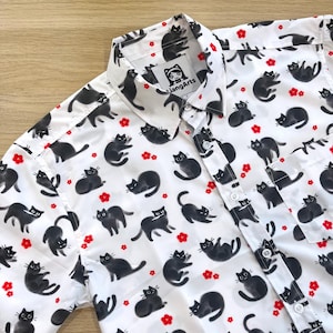 May include: White button-down shirt with a black cat print and red flower accents. The shirt has a collar and short sleeves. The brand name "XiangArts" is visible on the label.