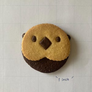 May include: A round cookie shaped like an otter face, featuring a light tan upper section and a dark brown lower section. The face has two small dark brown eyes and a dark brown diamond-shaped nose. A handwritten note indicates a size of 1 inch.