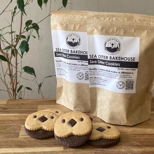 May include: Two tan paper pouches labeled Sea Otter Bakehouse Sea Otter Cookies with a net weight of 6.5 oz. In front, three round cookies feature a light tan top half with two small dark chocolate eyes and a dark chocolate nose, and a dark base.