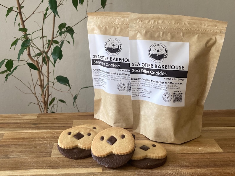 May include: Two tan paper pouches labeled Sea Otter Bakehouse Sea Otter Cookies with a net weight of 6.5 oz. In front, three round cookies feature a light tan top half with two small dark chocolate eyes and a dark chocolate nose, and a dark base.