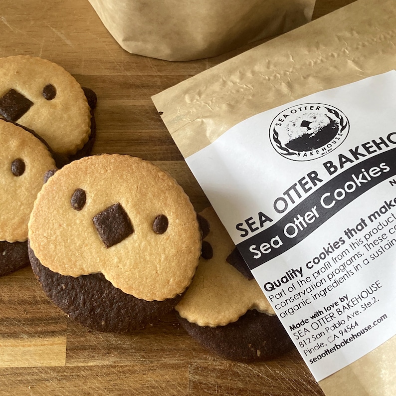 May include: Round cookies featuring a light tan top half and a dark brown bottom half, with two small dark dots for eyes and a dark square for a nose. Next to the cookies is a tan paper bag with a label reading SEA OTTER BAKEHOUSE Sea Otter Cookies.