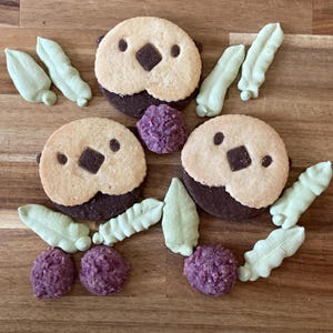 May include: Three round cookies shaped like sea otters with light tan faces and dark brown bodies, featuring dark chocolate noses and eyes. The cookies are surrounded by pale green leaf-shaped meringue pieces and small, textured purple berry-like treats.