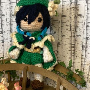 Puede incluir: Muñeca de ganchillo hecha a mano con un personaje de pelo azul oscuro, sombrero verde y atuendo verde y blanco. La muñeca está sobre un pequeño puente de madera con vegetación y flores artificiales. La muñeca tiene ojos negros y cara color beige claro.