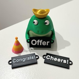 May include: A green frog figurine with a gold hat and holding a sign that says "Offer". Additional signs read "Congrats!" and "Cheers!". A party hat with orange and yellow stripes is also present.