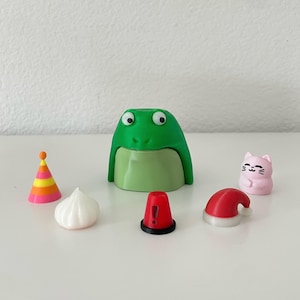 May include: A collection of small, colorful figurines and accessories. Includes a green frog-shaped container, a party hat, a white garlic-shaped item, a red cone with an exclamation point, a Santa hat, and a pink cat figurine.