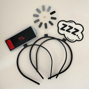 May include: A collection of black headbands with various attached novelty cutouts. Included are a battery icon showing 1%, a loading symbol, and a speech bubble with the word "ZZZ".
