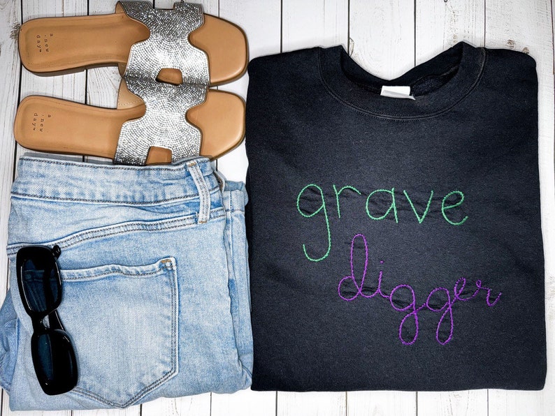 Pu&ograve; includere: Felpa nera con il testo "grave digger" ricamato in filo verde e viola.
