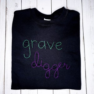 Pu&ograve; includere: Felpa nera con le parole "grave digger" ricamate in filo verde e viola.