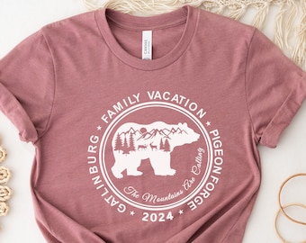 Puede incluir: Una camiseta color marrón rojizo con un gráfico circular blanco que presenta un oso, montañas y árboles. El texto alrededor del gráfico dice "Family Vacation * Pigeon Forge * Gatlinburg * The Mountains Are Calling * 2024".