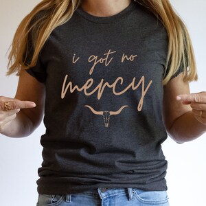 May include: A dark gray t-shirt with the text "i got no mercy" in a light brown color. A stylized cow skull is below the text.
