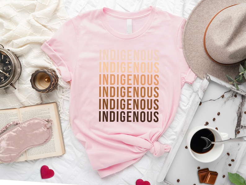 Może przedstawiać: R&oacute;żowy T-shirt z napisem "Indigenous" powt&oacute;rzonym sześć razy w gradiencie brązowych odcieni.