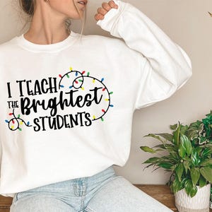 May include: White sweatshirt with the text "I Teach the Brightest Students" in black, with colorful string lights. The sweatshirt is long-sleeved and has a crew neck. The design is centered on the front.