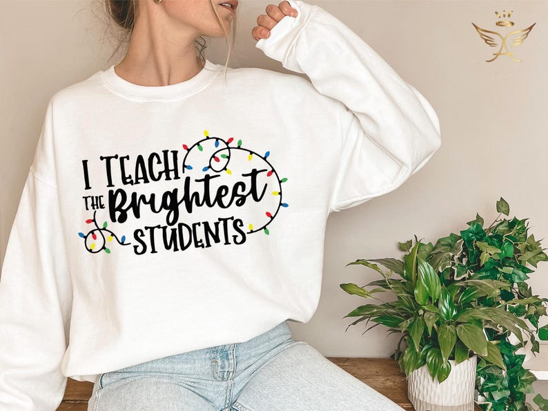 May include: White sweatshirt with the text "I Teach the Brightest Students" in black, with colorful string lights. The sweatshirt is long-sleeved and has a crew neck. The design is centered on the front.