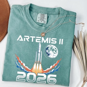 May include: A teal-colored t-shirt with the words "ARTEMIS II" and the year "2026" printed in white. The shirt features a graphic of a rocket launching into space, with a moon illustration. A gold necklace rests on the shirt.