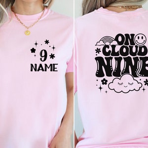 May include: Pink t-shirt with a black graphic on the front that says "*9* NAME" and a black graphic on the back that says "ON CLOUD NINE" with a cloud, rainbow, and stars.