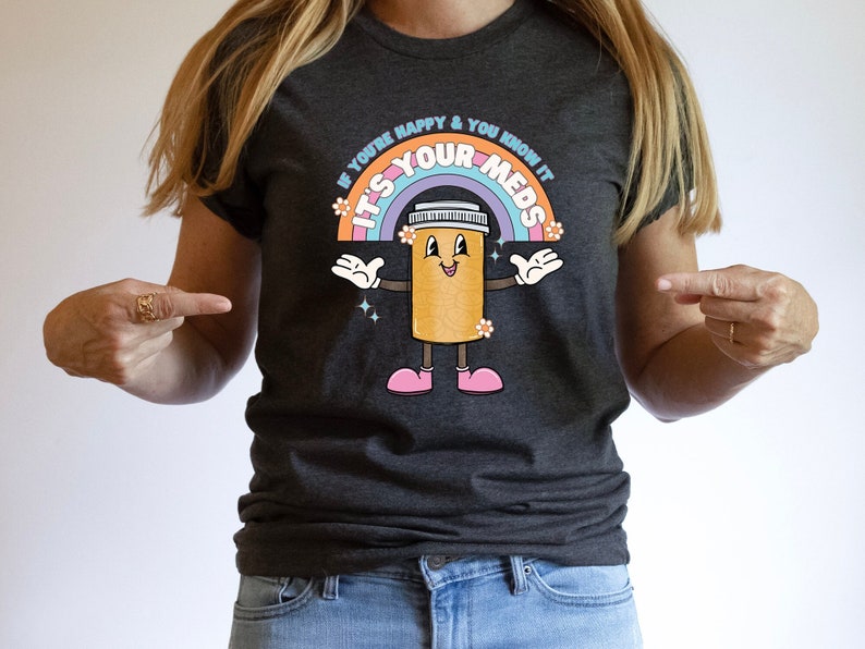 Peut inclure: Un t-shirt gris fonc&eacute; avec un arc-en-ciel et une pilule de dessin anim&eacute; avec un visage souriant. Le texte sur le t-shirt est "Si tu es heureux et que tu le sais, c'est tes m&eacute;dicaments."