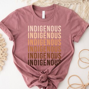 Może przedstawiać: Koszulka w kolorze malwy z napisem "Indigenous" powt&oacute;rzonym sześć razy w gradiencie brązowych odcieni.