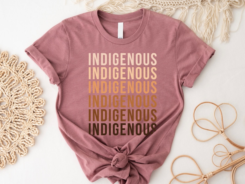 Może przedstawiać: Koszulka w kolorze malwy z napisem "Indigenous" powt&oacute;rzonym sześć razy w gradiencie brązowych odcieni.