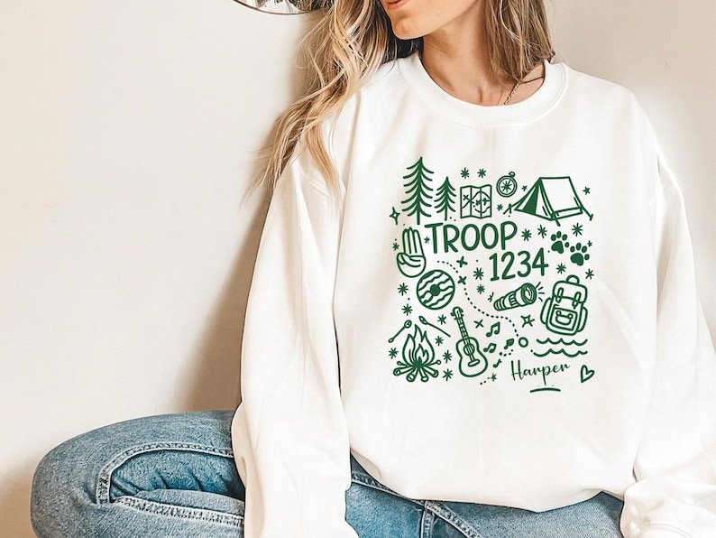 May include: White sweatshirt with a green graphic design featuring camping-themed illustrations and the text "TROOP 1234" and "Harper". The design includes trees, a tent, a compass, a guitar, and a backpack.