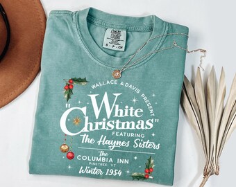 May include: A teal-colored t-shirt with the text "White Christmas" in a vintage-style design. The shirt features holly and ornaments, with the text "The Haynes Sisters" and "Winter 1954". A gold necklace and a brown hat are also visible.