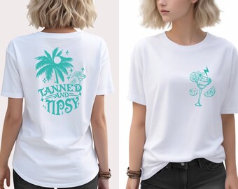 May include: White t-shirt with a teal green design on the back. The design features a palm tree, a cocktail, and the text "Tanned and Tipsy". The front of the shirt has a teal green design of a cocktail with a lime wedge.