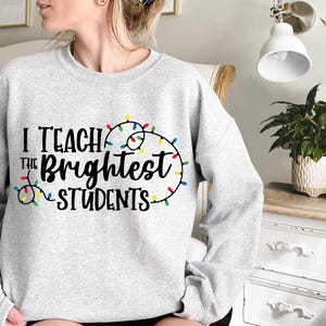 May include: A light gray sweatshirt with the text "I Teach the Brightest Students" in black script. The text is accented with a string of colorful Christmas lights. A comfortable, casual top for teachers.