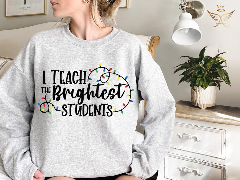 May include: A light gray sweatshirt with the text "I Teach the Brightest Students" in black script. The text is accented with a string of colorful Christmas lights. A comfortable, casual top for teachers.