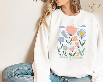 May include: White sweatshirt featuring a floral design with pink, blue, and yellow flowers, and green stems. The text "CONSIDER HOW THE WILDFLOWERS GROW" is printed below the flowers. The sweatshirt is a comfortable, casual style.